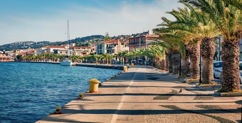 A wide-angle view of the Argostoli waterfront in Kefalonia, Greece, with fishing boats docked at the stone quay and a palm-lined promenade against a backdrop of mountains.
