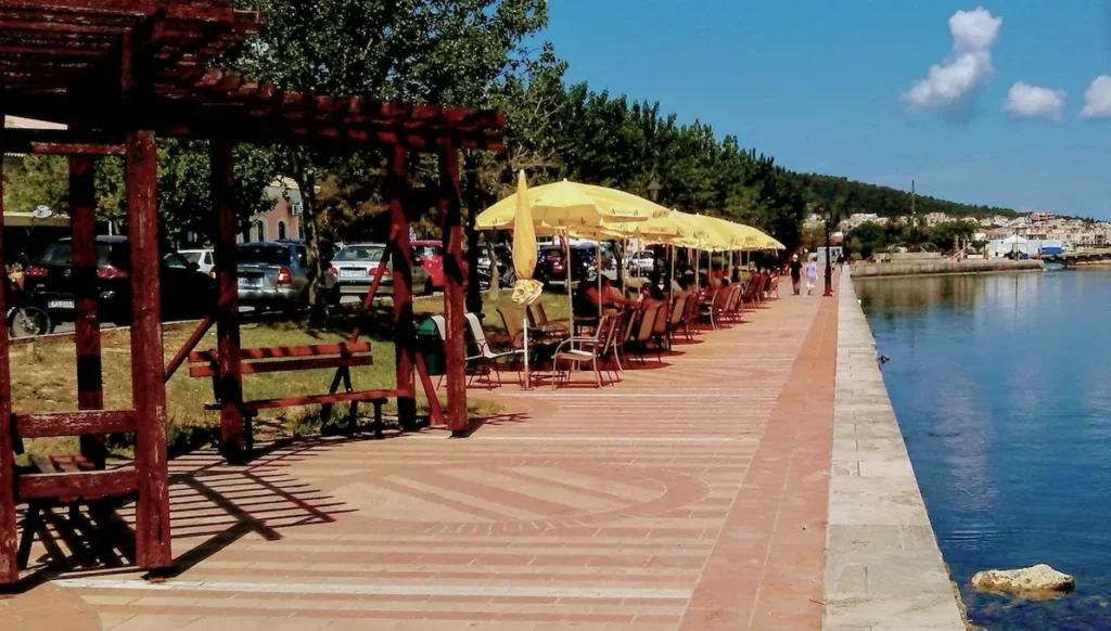 A wide view of the paved pedestrian promenade in Argostoli, lined with palm trees and streetlamps, stretching along the calm harbor at dusk.