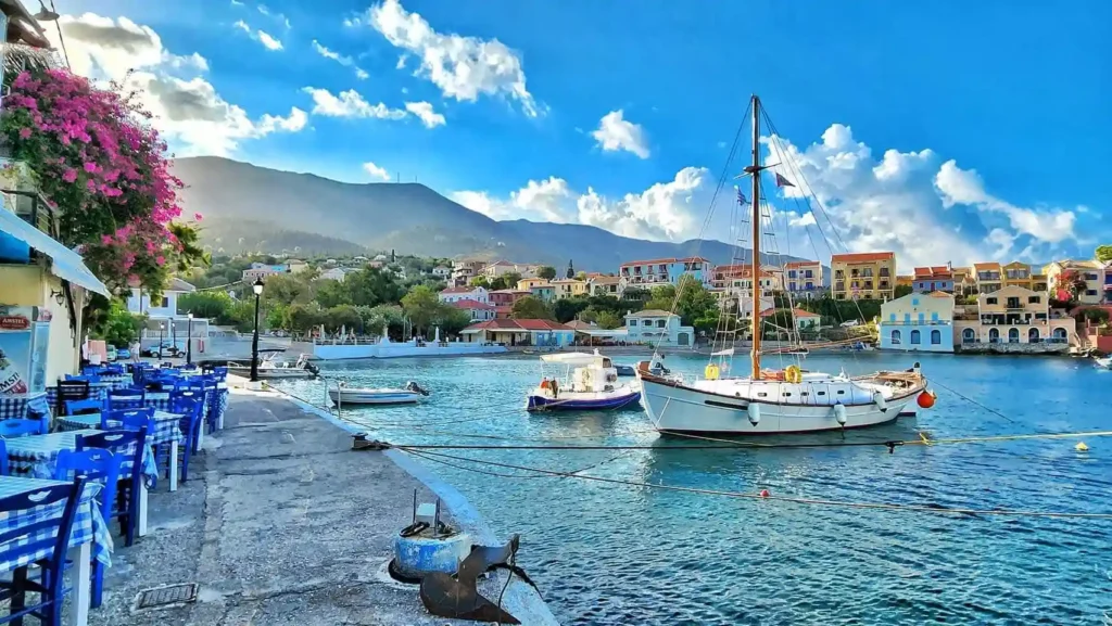 A view of colorful wooden fishing boats moored in the turquoise waters of Assos harbor, with a blue-clothed taverna table in the foreground.