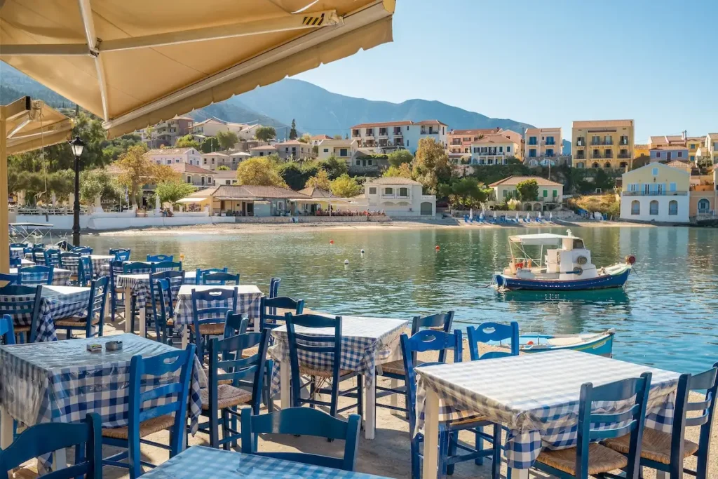 A traditional Greek taverna with wooden tables and chairs set on the stone quay of the Assos harbor, with the village’s colorful houses in the background.