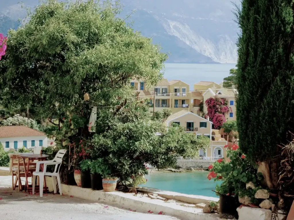 A framed view through Mediterranean greenery looking down from a high hillside toward the turquoise bay and colorful village of Assos.
