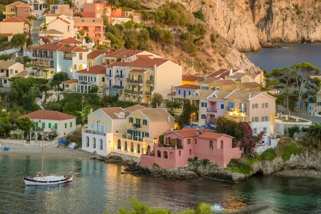 Aerial perspective of the small coastal village of Assos, Kefalonia, with traditional houses in shades of beige, garnet (grenade), and light yellow, built on a thin strip of land leading to a Venetian fortress.