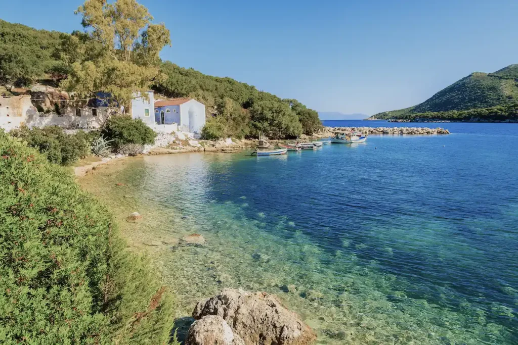 A calm coastal bay in Kefalonia featuring a small, light blue Greek Orthodox church and a few colorful wooden fishing boats moored in clear water.