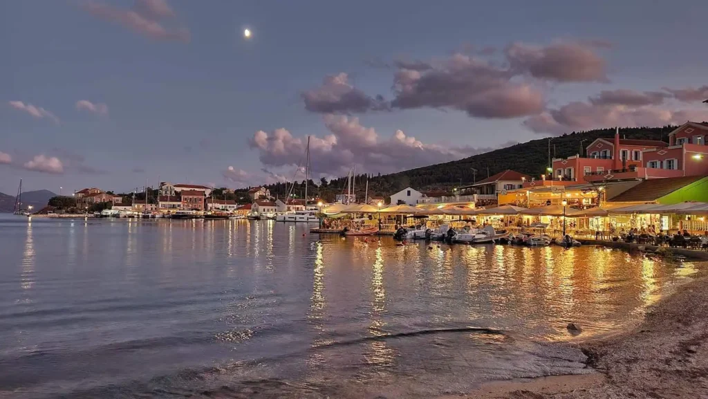 A view of the historic Fiscardo waterfront at sunset, with traditional neoclassical buildings reflected in the calm harbor waters.
