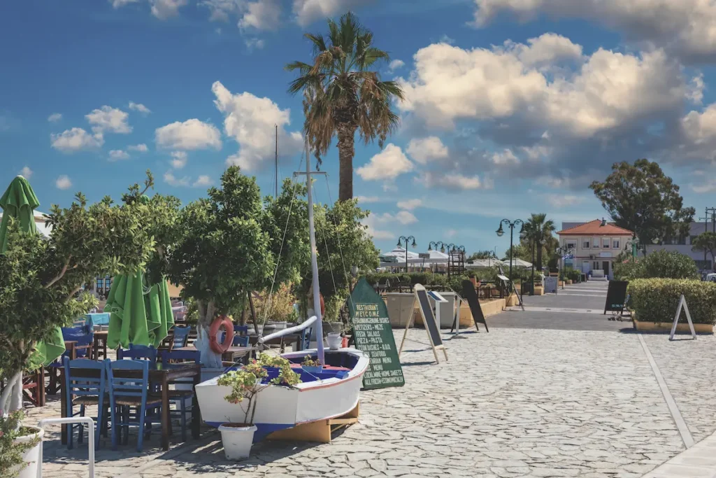 A wide town square in Lixouri, Kefalonia, with outdoor taverna tables under large trees, traditional buildings, and locals gathered to eat and socialize.
