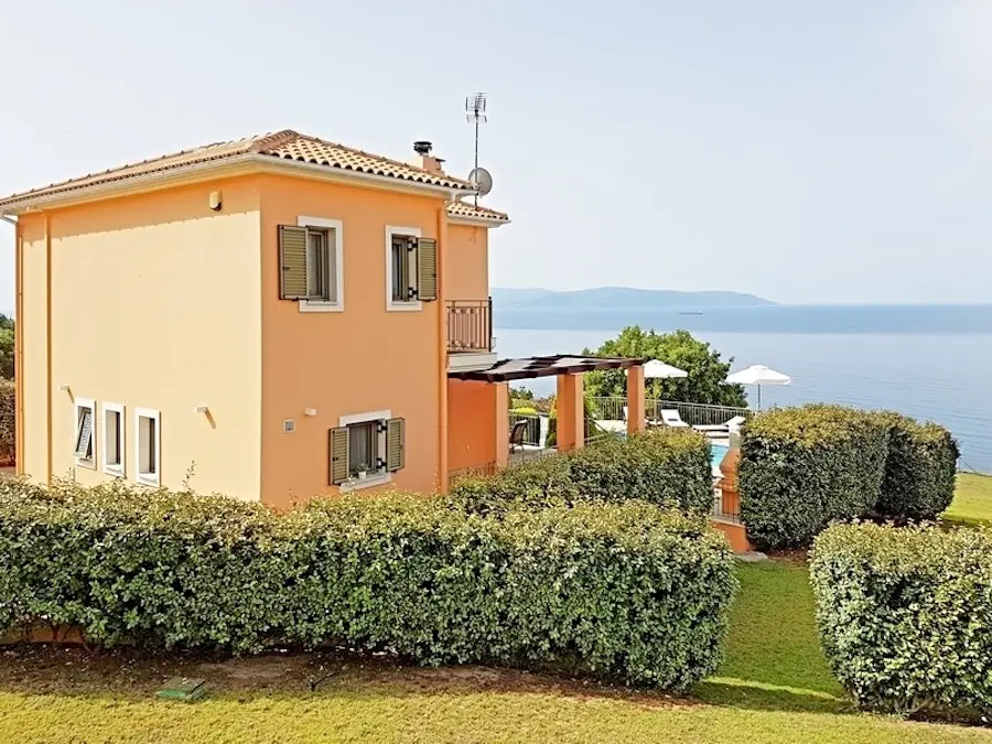 A grand, two-story neoclassical mansion painted in ochre-orange with white architectural details and a wide view of the Ionian Sea.