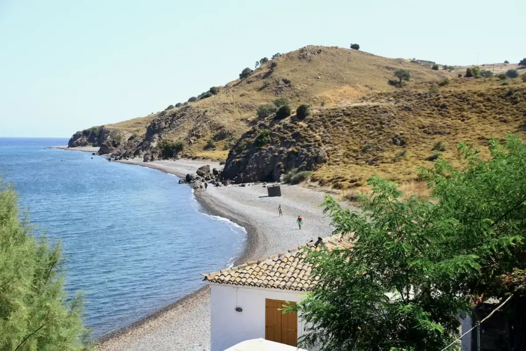 A wide, natural shoreline of smooth dark pebbles meeting the deep blue of the Aegean.