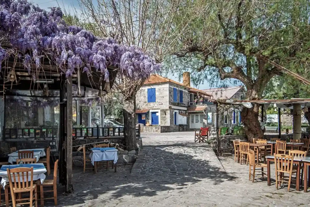 A village square featuring traditional stone architecture, the outdoor seating of a taverna, and a prominent stone house.