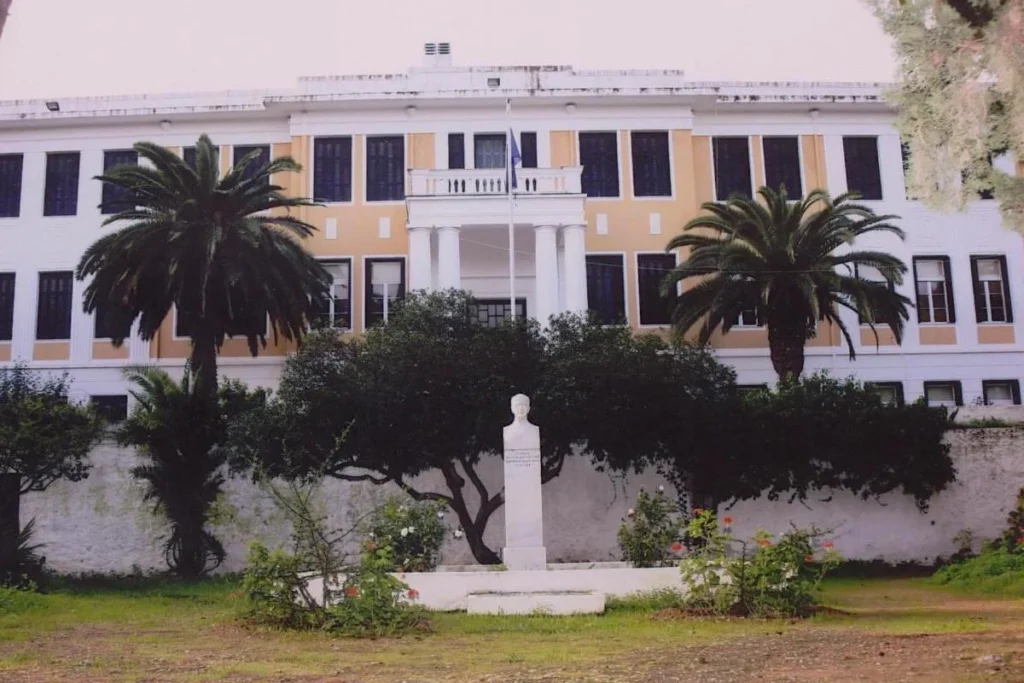 The grand neoclassical facade of the Anargyreios and Korgialeneios School, surrounded by pine trees with a view of the Saronic Gulf.