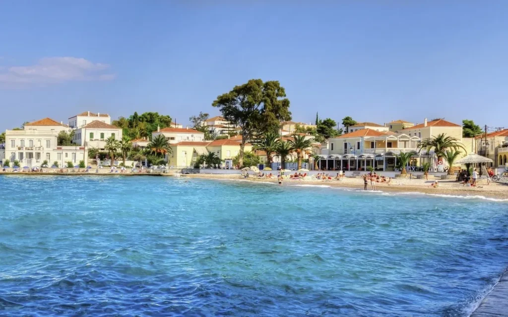 A wide panoramic shot of a pebbled beach on Spetses with rows of sunbeds and blue umbrellas under a clear sky.
