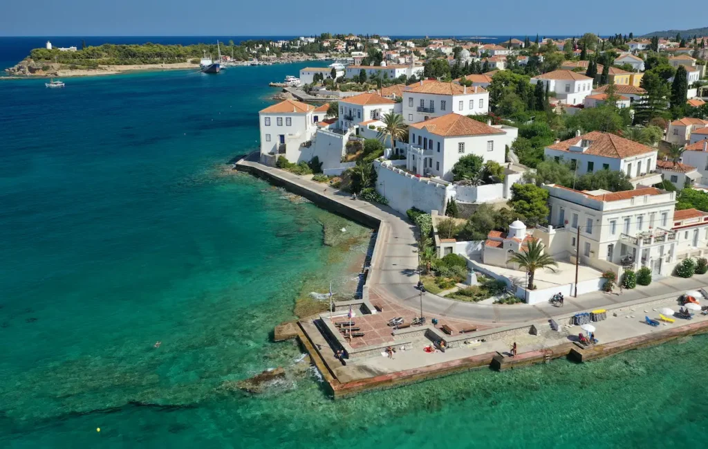 A high-angle aerial view of the white-domed Agios Mamas chapel surrounded by the terracotta-tiled roofs of traditional sea-captain mansions on Spetses.