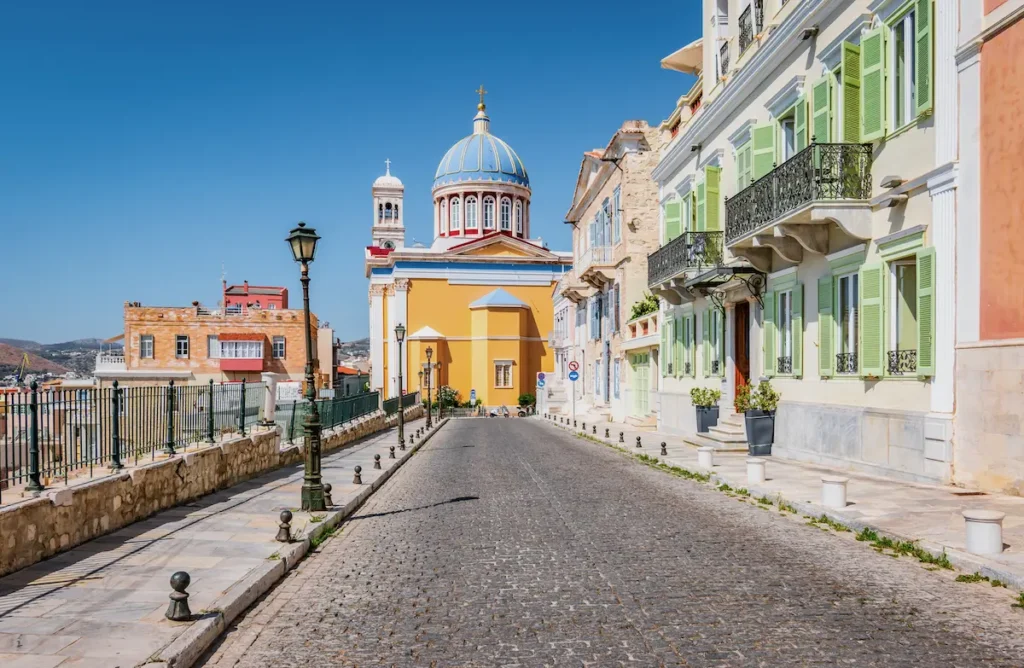 A paved street in Ermoupoli with colorful houses leading toward the blue dome of St. Nicholas Church.