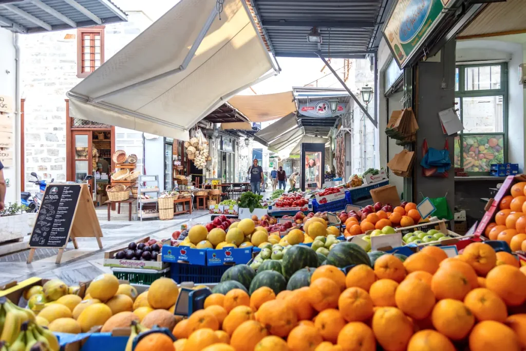 A vibrant outdoor fruit and vegetable market on a marble-paved pedestrian street in Ermoupoli, Syros, near the central square.