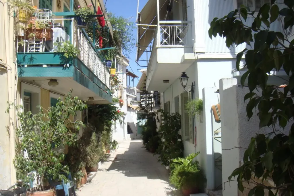 A narrow stone‑paved residential alleyway (sokaki) in Lefkada town, filled with potted green plants and jasmine vines.