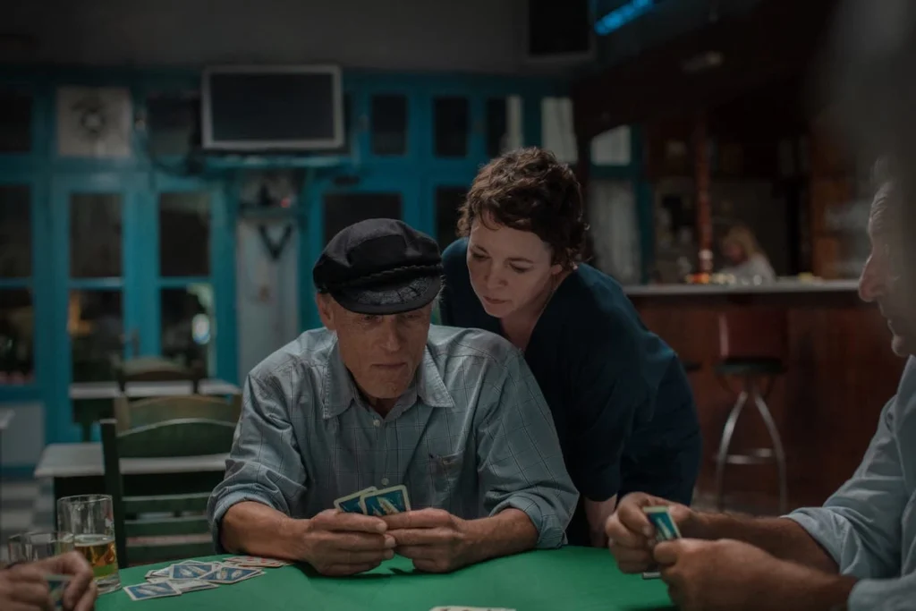 Ed Harris as Lyle playing cards at a wooden table while Olivia Colman watches him in a traditional Greek interior.