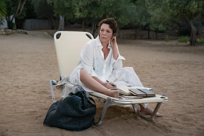 Actress Olivia Colman as Leda sitting on a sunbed under a beach umbrella on a pebbled Greek beach.
