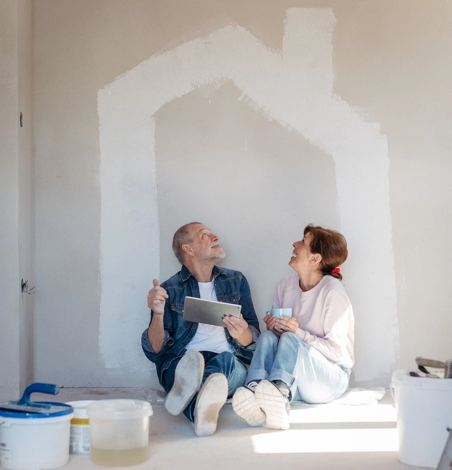 A senior man and woman sitting on the floor of a sunlit room during a home renovation, surrounded by tools and construction materials.