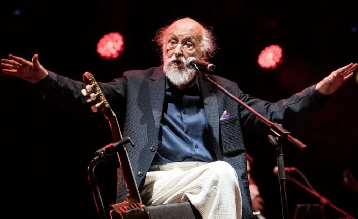 A candid shot of Greek singer-songwriter Dionyssis Savvopoulos performing on stage, gesturing expressively while sharing a story with the audience.