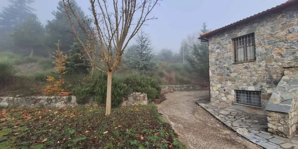 A traditional old stone house sits nestled in a mountain village in the Feneos Region of Greece, enveloped in thick winter fog, offering a quiet, remote atmosphere.