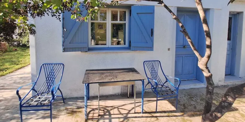 A charming Greek summerhouse with light blue doors and windows, a large green garden, and metal chairs, representing affordable real estate in Greece.