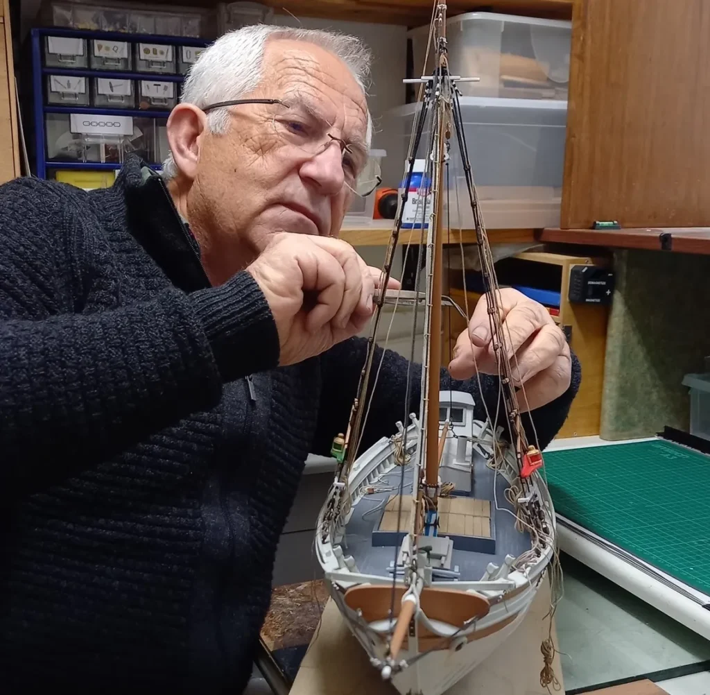 Vangelis Koumarianos leaning over a workbench, carefully assembling a detailed wooden model boat in his Nafplio home.
