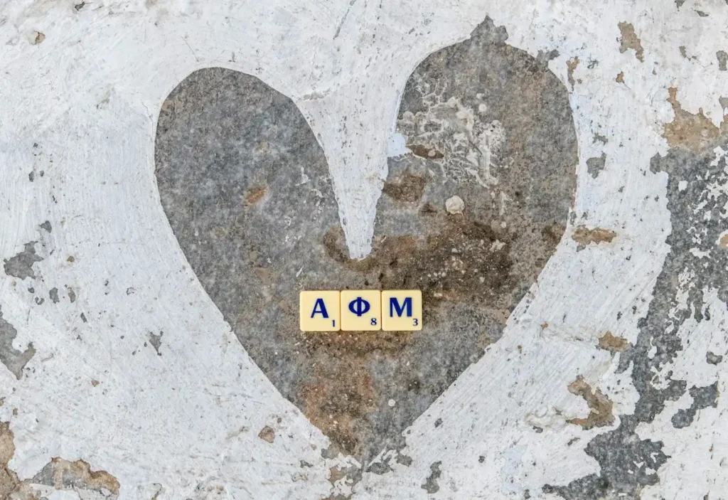 A ground-level photograph capturing the Greek letters ΑΦΜ (Tax ID Number) and a hand-painted white heart on a traditional stone-paved street. The pavement features typical white-painted outlines.