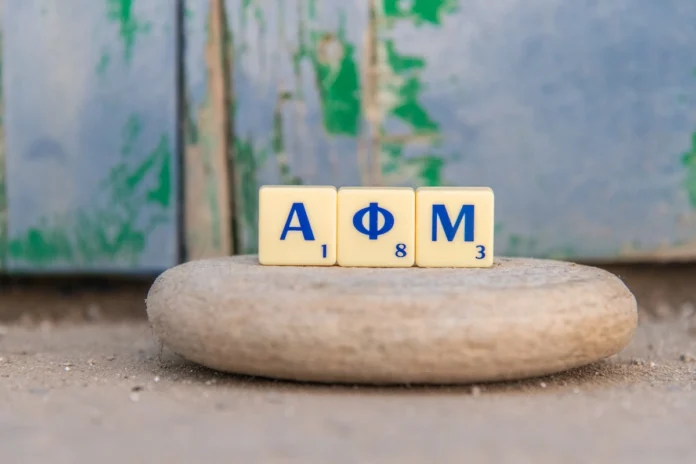 Scrabble letters spelling in greek AFM are arranged in the foreground against a sunlit Greek backdrop.