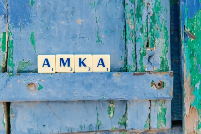 Scrabble tiles spelling AMKA positioned in the foreground with a blurred Greek coastal background.