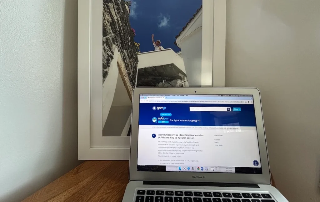 Laptop showing gov.gr tax portal with a blurred background of a woman waving from a traditional Greek balcony.