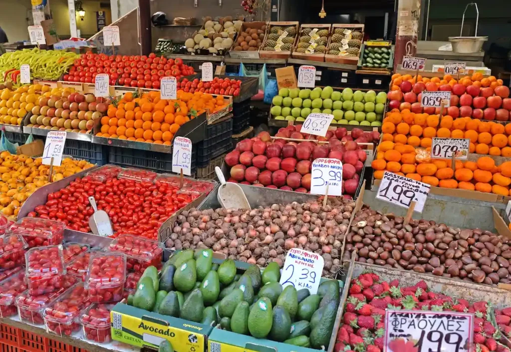A close-up, top-down view of two large, heavy bags filled with tomatoes, cucumbers, greens, and seasonal fruits resting on a stone pavement.