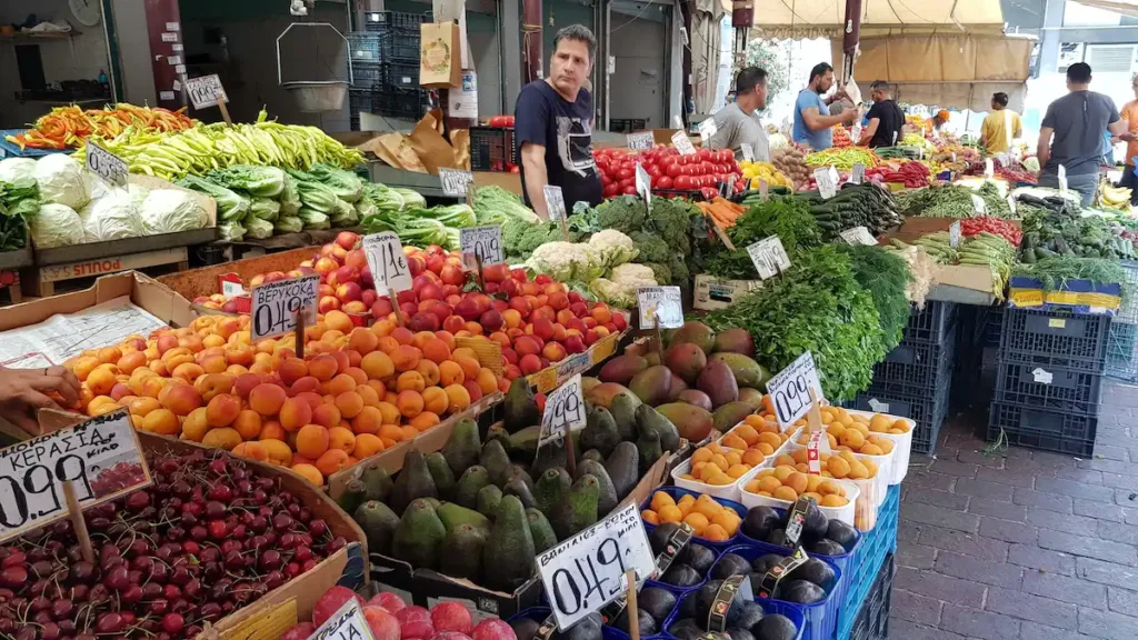 A friendly local vendor standing behind a wooden market stall overflowing with fresh oranges and lemons, with hand-written price tags visible.