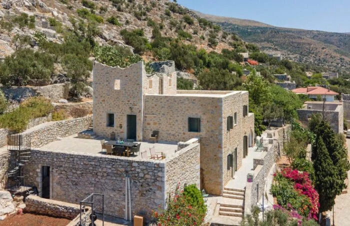 Aerial view of a traditional stone tower house in Mani, Peloponnese, surrounded by a dry, rugged landscape under a bright, burning sun.