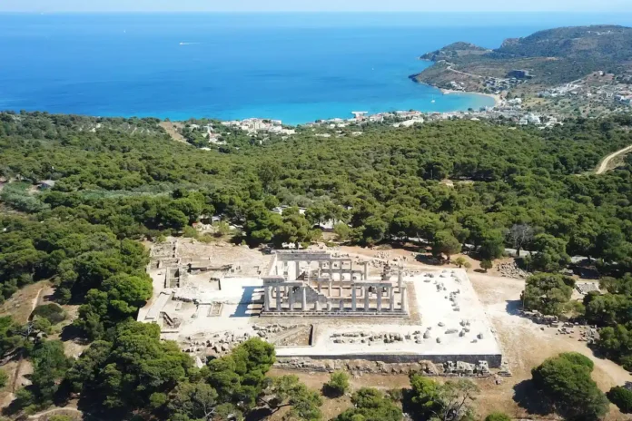 Wide aerial shot of the Temple of Aphaia surrounded by pine and pistachio trees with the blue Saronic Gulf in the distance.