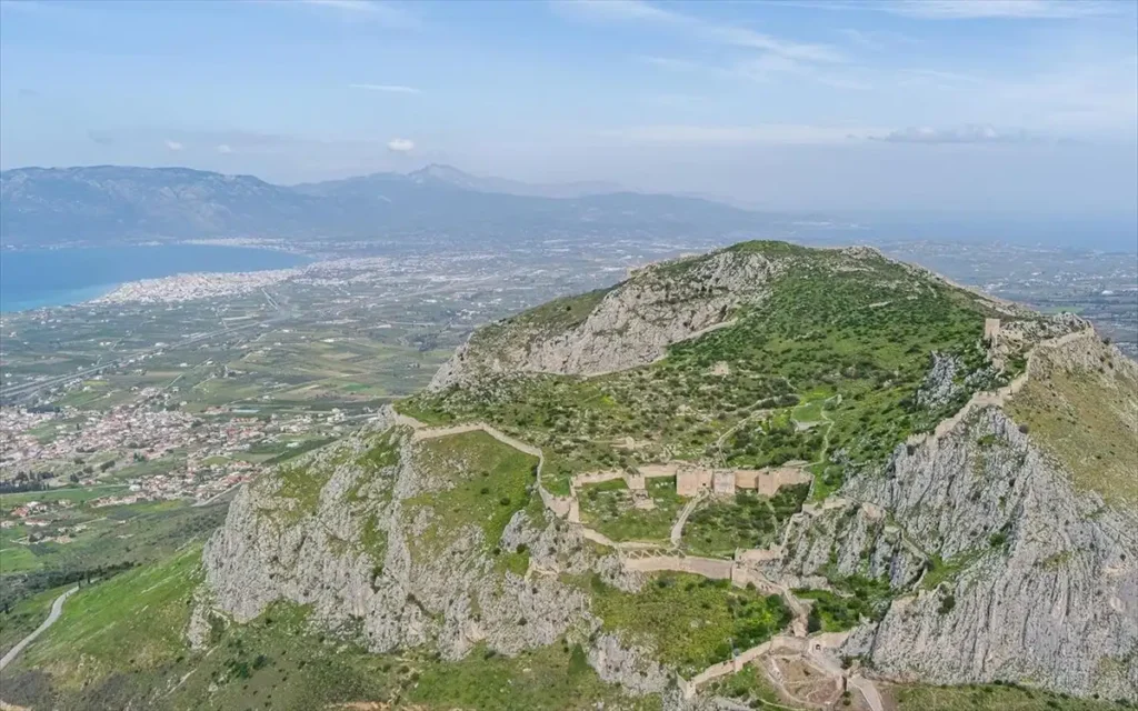 Aerial view showing the fortified summit of Akrokorinthos with its stone walls and ruins, overlooking the blue waters of the Corinthian Gulf and the town of Loutraki in the distance.
