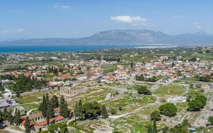 Aerial view of Ancient Corinth village showing the proximity of traditional houses to the Temple of Apollo and the blue coastline of the Corinthian Gulf in the distance.