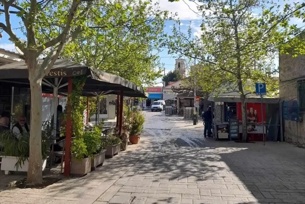 A traditional paved stone street in the village of Ancient Corinth lined with low-rise buildings and greenery.