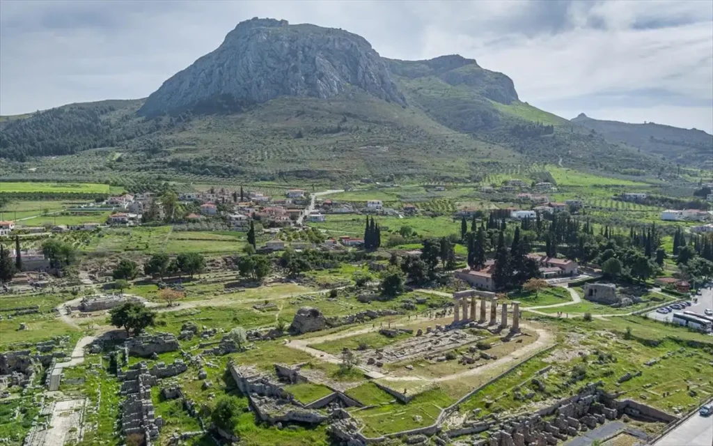 A wide landscape shot encompassing the Temple of Apollo, the modern village houses, and the Akrokorinthos mountain in a single vertical perspective.
