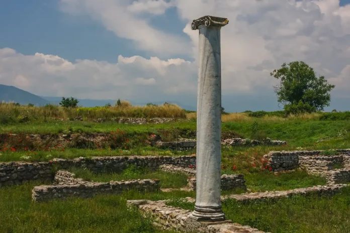 A solitary ancient stone column standing tall amidst low foundation ruins and wildflowers.