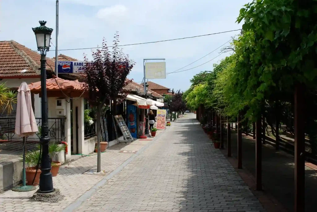 A wide, stone-paved pedestrian street in modern Dion Village with local shops and cafe seating.