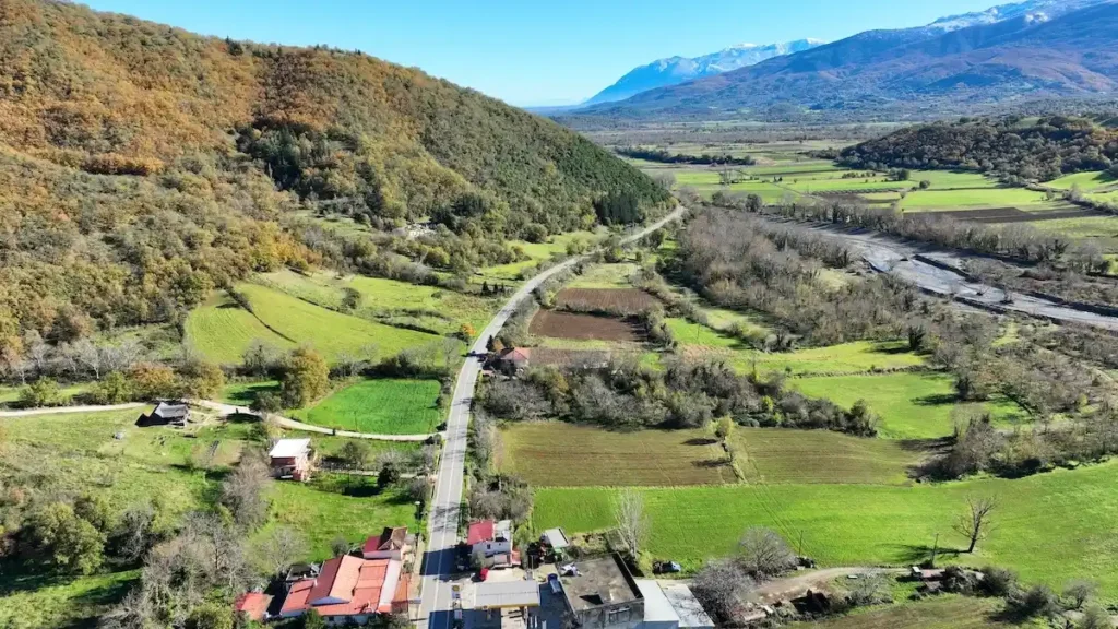 A wide view of a lush green valley in Karpenissi with scattered traditional houses at the foot of massive, towering mountains, featuring early autumn orange foliage.