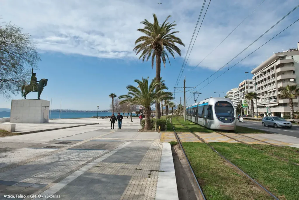 Wide shot of the paved coastal promenade in Palaio Faliro with residents walking and cycling under the Greek sun.