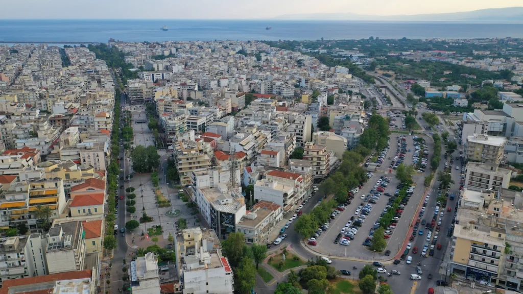 An aerial panorama of Kalamata City, showing the dense grid of low-rise buildings interspersed with numerous parks, squares, and tree-lined avenues, stretching toward the sea.