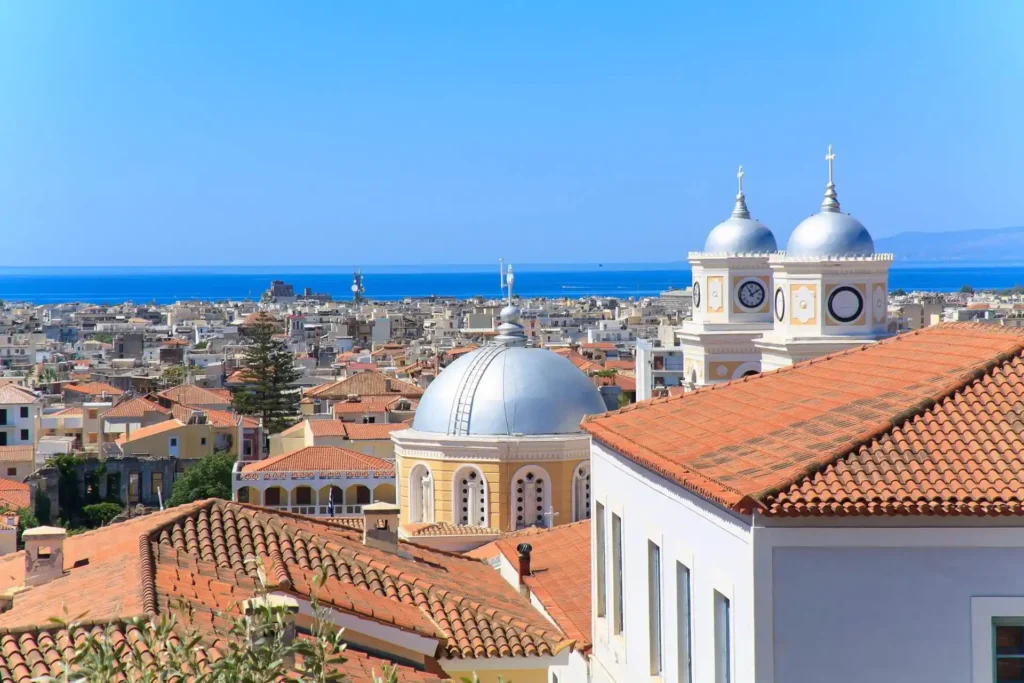 A high-angle view looking across the red-tiled roofs of the Kalamata historical center (Chora), culminating in the prominent, vibrant blue domes and stone belfry of the Holy Apostles Church.