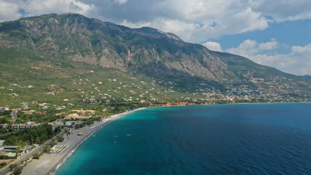 A wide view of the long, sandy Kalamata beach with small waves, looking toward the massive, dark green peaks of Mount Taygetos in the background.
