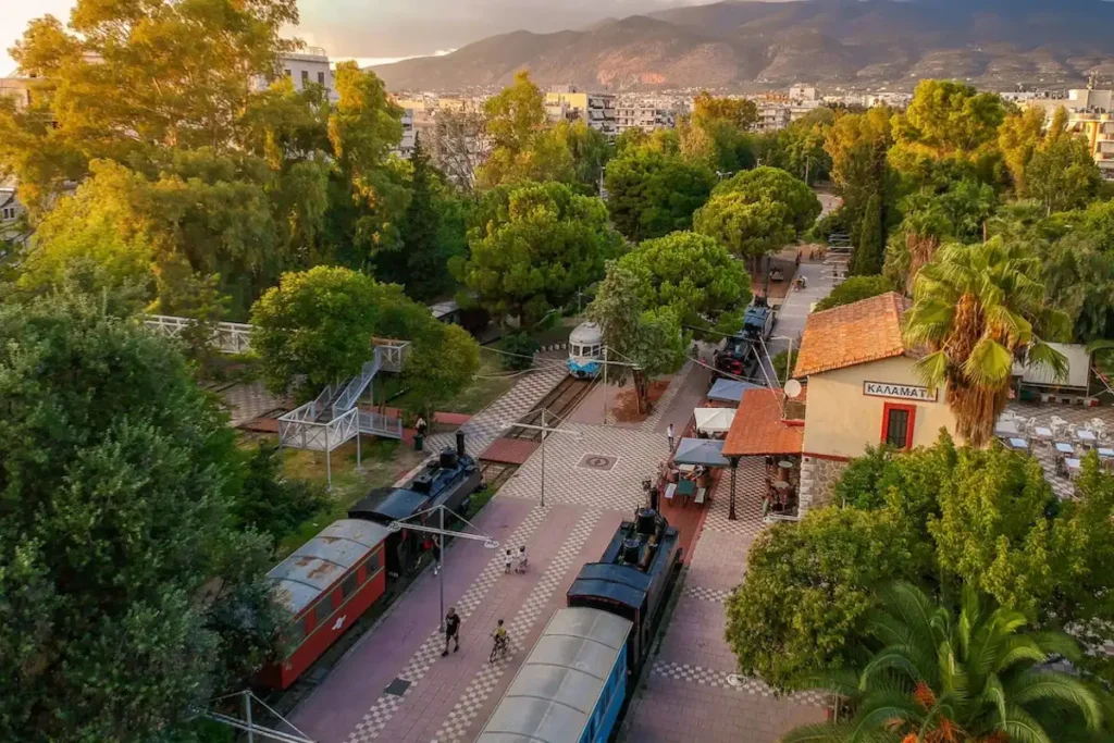 A view of the historic Kalamata OSE Railway Park, showing a wide paved walking path with a vintage train car on a parallel track, bordered by green trees.