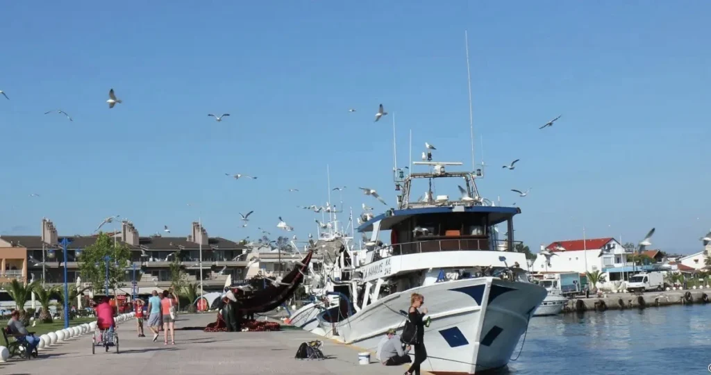 A wide-angle view of the Keramoti harbor promenade, showing colorful fishing boats, a large ferry boat, and leisure yachts docked along the paved walkway, with people strolling past cafes.