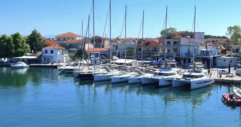 An aerial view of Keramoti harbor showing modern yachts and catamarans docked near a small, traditional Greek fish tavern at the water's edge.