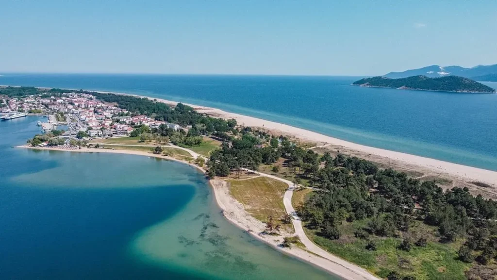A high-altitude aerial photograph of Keramoti, Greece, showing the urban layout of the town bordering the golden sands and green pine groves of the Ammoglossa peninsula.