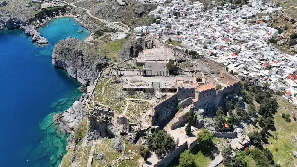 An aerial drone view of a historic Greek coastal settlement, featuring a hilltop ancient citadel overlooking a traditional whitewashed village and a turquoise bay.