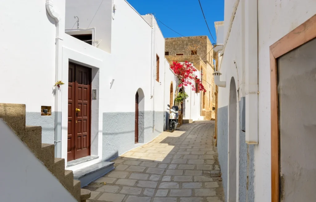 A narrow, traditional cobblestone street winding between white stone walls in Lindos village.